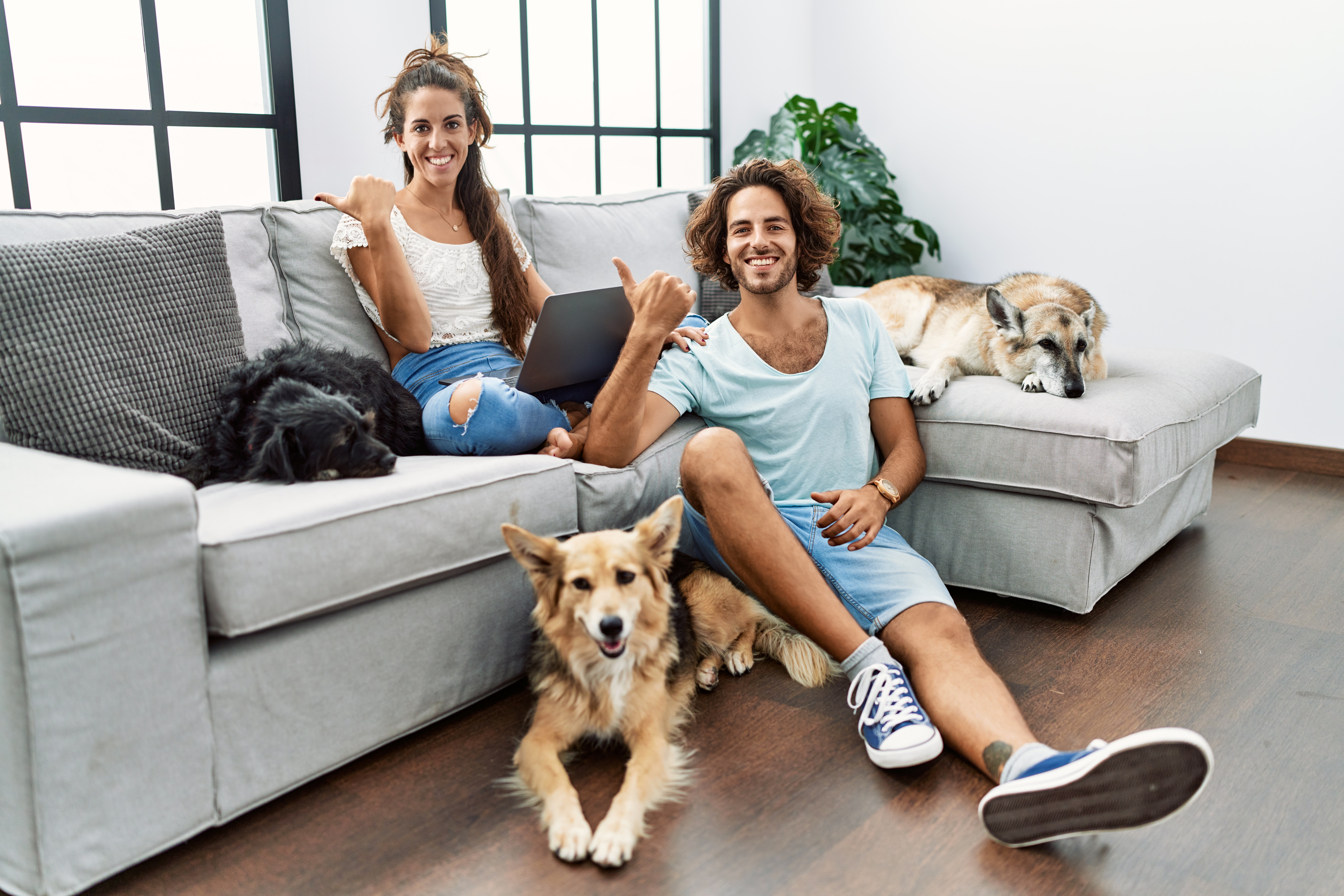Young hispanic couple with dogs relaxing at home pointing to the back behind with hand and thumbs up, smiling confident